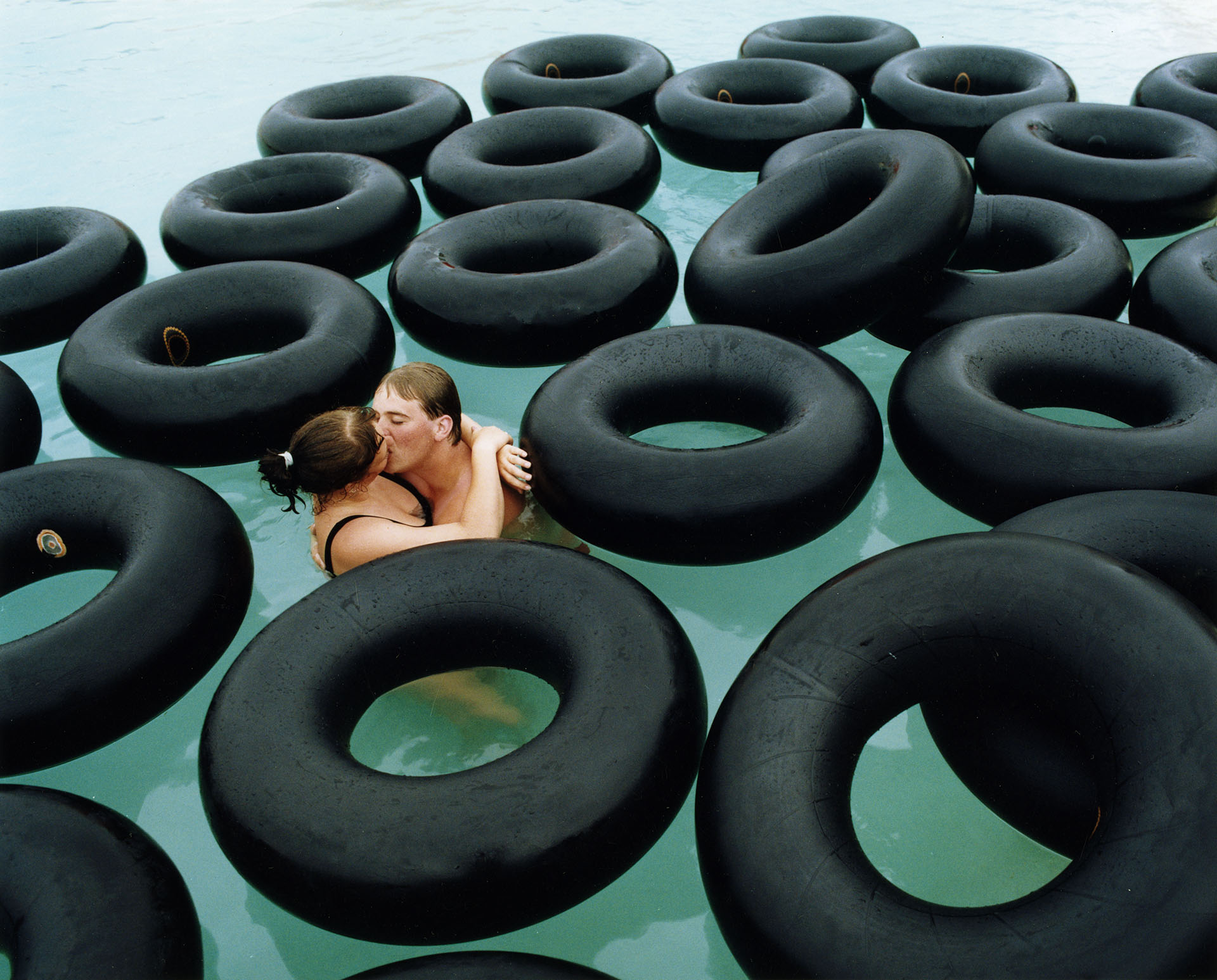 Ett par som kysser varandra i en pool med grönt vatten omringade av svarta däck.