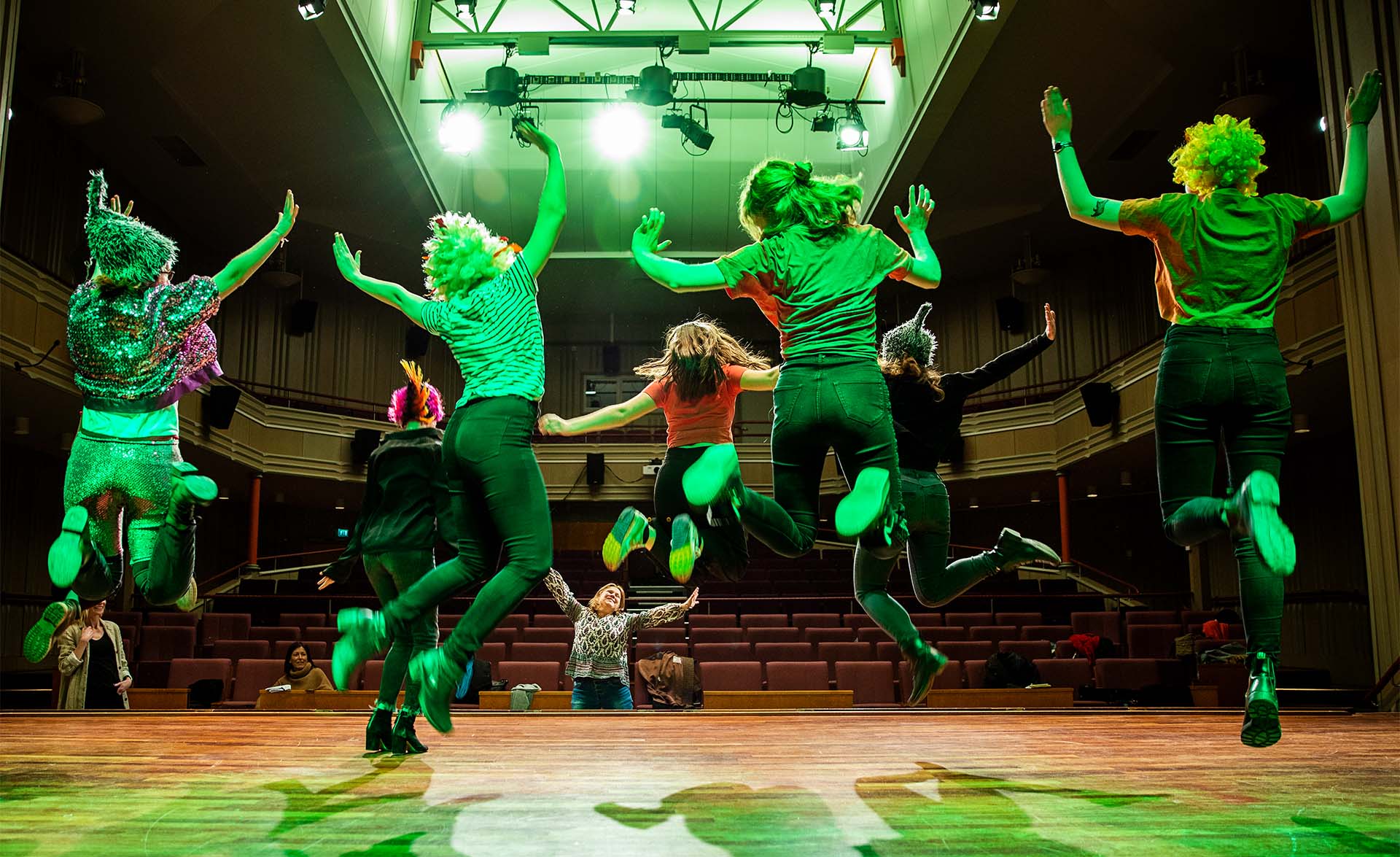 En musikalgrupp gör ett hopp upp i luften samtidigt på en scen i starkt grönt scenljus.